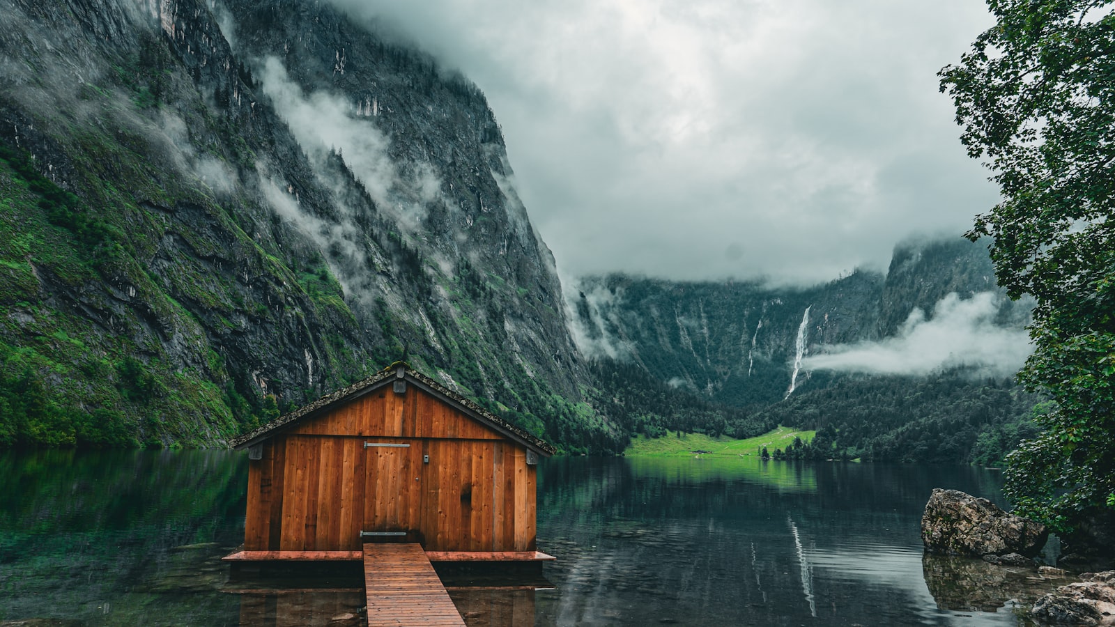 A wooden lakeside cabin surrounded by fog-wrapped mountains
